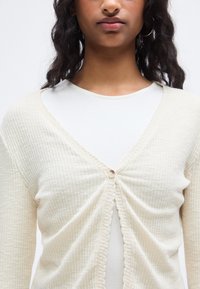 Ανοιχτό μπεζ ζακέτα με κουμπιά και ριμπ υφή, με κυματιστό τελείωμα, φορεμένη πάνω από λευκό τοπ με στρογγυλή λαιμόκοψη. Κολακευτικό V-λαιμόκοψη σχέδιο.