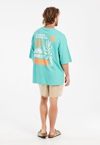 Tyrkysové oversized tričko s grafickými motivy v oranžové a bílé barvě, krátké rukávy a pohodlný střih, v kombinaci s béžovými šortkami.