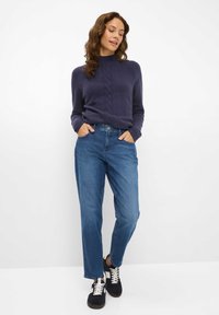 Maglione lavorato a maglia blu navy con collo alto, caratterizzato da un motivo testurizzato. Jeans dritti azzurri, abbinati a sneakers nere con lacci bianchi.