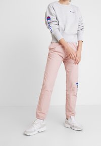 Un sweatshirt gris avec des logos bleus et rouges, assorti à un pantalon taper léger rose. Des baskets blanches avec des accents texturés complètent la tenue.