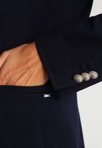 Giacca blu navy in tessuto liscio, con una tasca prominente e tre bottoni decorativi argentati sul polsino. Dettagli di design sartoriali visibili.