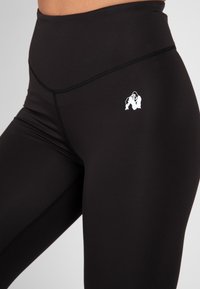 Zwarte legging gemaakt van rekbaar stof, met een hoge tailleband en een klein wit gorilla-logo aan de linkerkant. Gladde structuur, aansluitend ontwerp.