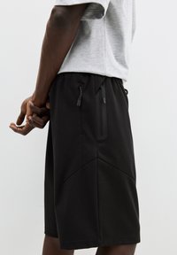 Pantaloni scurți atletici negri cu o textură netedă, talie elastică și detalii cu logo; au două buzunare laterale cu închidere cu șnur.