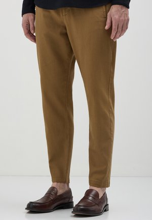Man wearing brown tapered pants and dark brown leather loafers, standing against a plain white background.