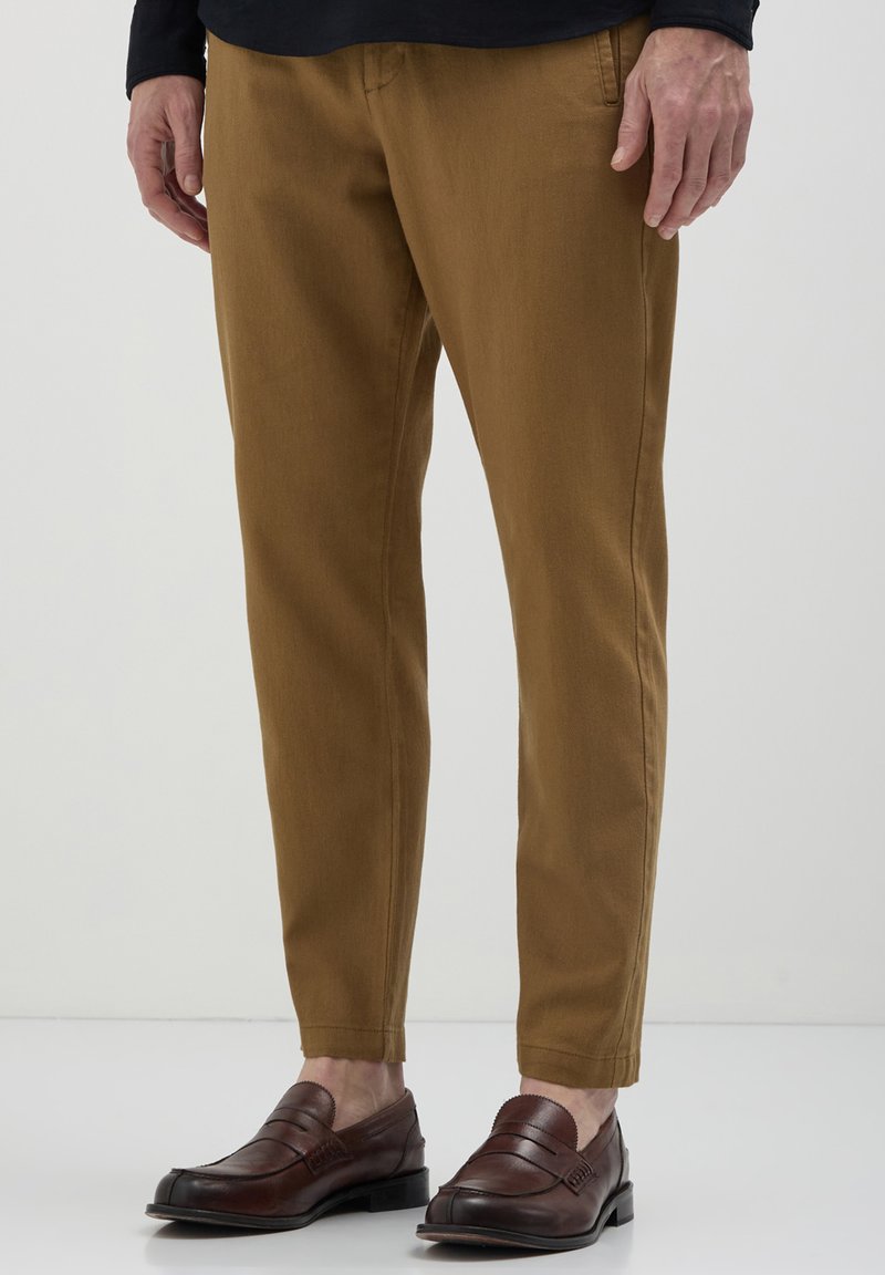 Homme portant un pantalon brun fuselé et des mocassins en cuir brun foncé, se tenant contre un fond blanc uni.