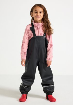 Zwarte waterdichte overalls met verstelbare banden, gecombineerd met een lichtroze fleecjas met dierenprints. Felrode rubberen laarzen.