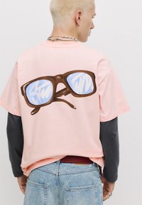 Tricou din bumbac roz cu mânecă scurtă, având un grafic mare maro cu ochelari pe spate, cu lentile reflectorizante albastre.