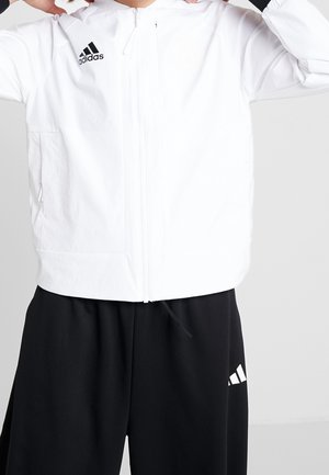 Osoba nosząca białą kurtkę adidas na suwak i czarne spodnie adidas, poprawia kołnierz kurtki obiema rękami.