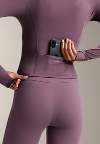 Fialová športová halenka s dlhými rukávmi a vreckom na smartfón na zadnej strane, v kombinácii s vyššími legínami. Elastická tkanina.