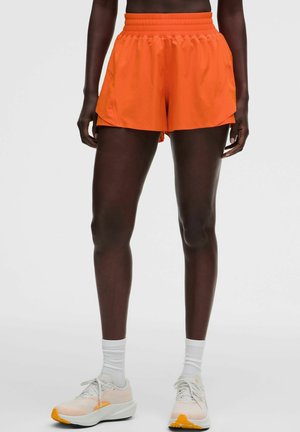 Persoon die feloranje sportbroekjes, witte enkelsokken en witte en oranje hardloopschoenen draagt, staand tegen een lichtgrijze achtergrond.