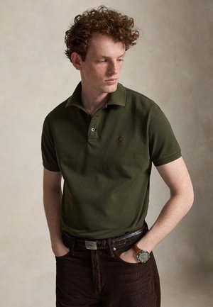Jeune homme aux cheveux roux bouclés portant un polo vert olive, un pantalon foncé et une montre-bracelet, regardant vers le bas et sur le côté.