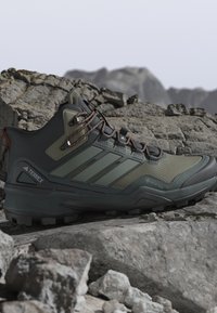 Zapatilla de senderismo Adidas Terrex Gore-Tex en verde oliva y negro apoyada sobre rocas grises y escarpadas de montaña.