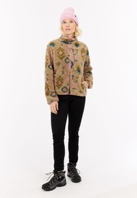 Woman wearing patterned beige fleece jacket, pink beanie, black pants, and hiking boots standing against a white background.