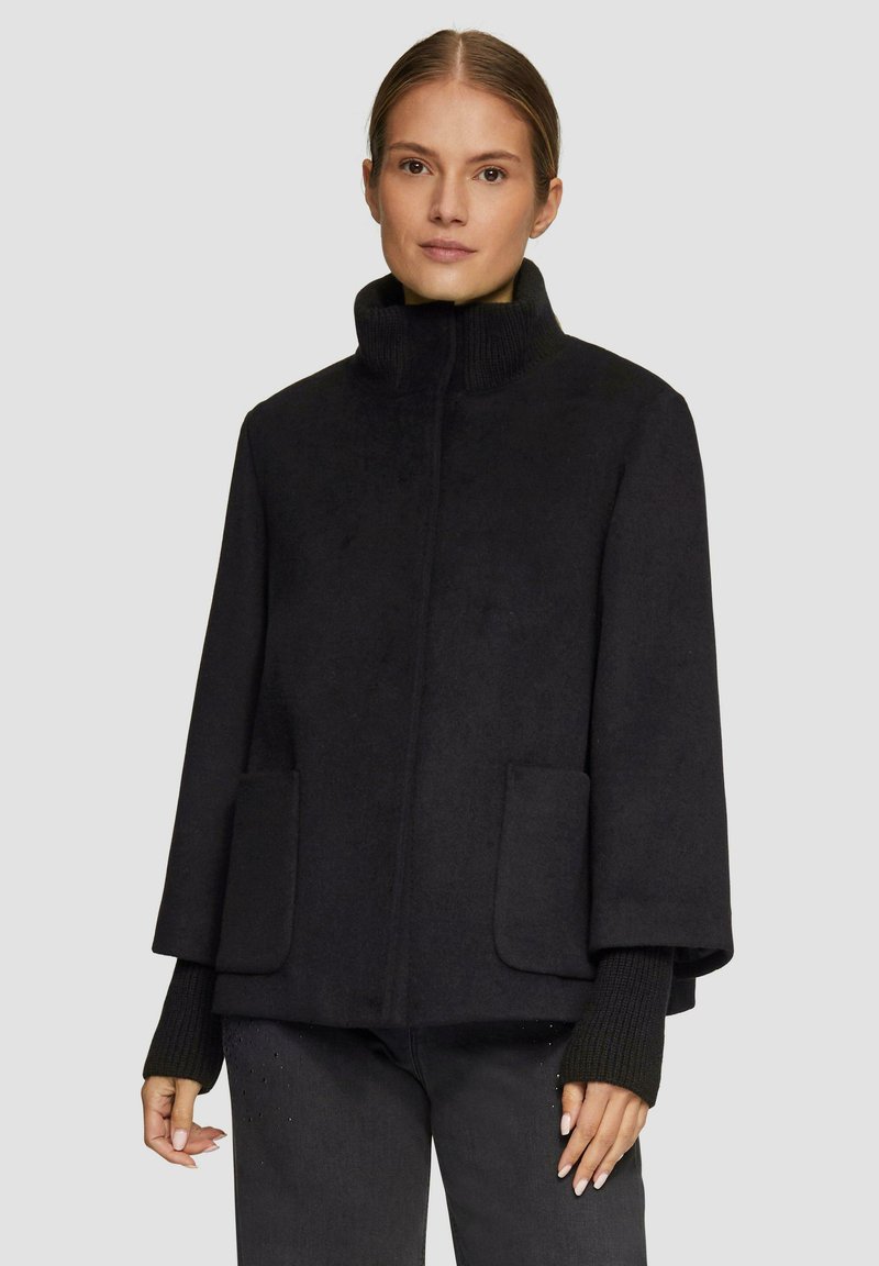 Czarny płaszcz z wełny z wysokim kołnierzem, bocznymi kieszeniami i detalami ściągaczy na rękawach. Materiał ma gładką teksturę i wyraźnie zarysowany kształt.