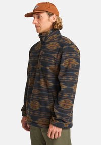 Pull en polaire bleu marine avec un motif géométrique en orange et marron. Présente un col montant et un design à demi-zip, avec des poches latérales.