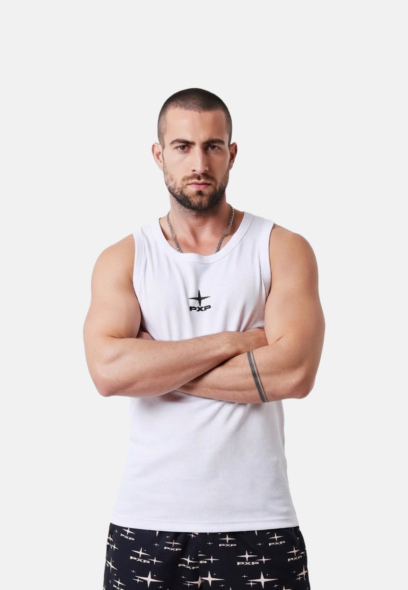 Homme aux cheveux courts et barbe, portant un débardeur blanc et un short noir à motifs d'étoiles, se tient les bras croisés devant un fond uni.
