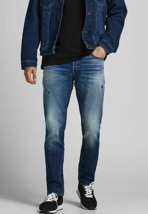 Mann trägt ausgewaschene blaue Slim-Fit-Jeans mit leichten Used-Effekten, kombiniert mit einer dunklen Jeansjacke, schwarzem Hemd und schwarzen Sneakern mit weißen Schnürsenkeln.