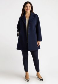 Marinblå ullkappa med slagkrage, två framfickor och silverknappar, tillsammans med marinblå slim-fit byxor och spetsiga metalliska klackar.
