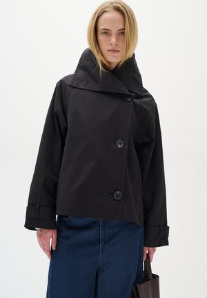 Cappotto corto nero con colletto alto, grandi bottoni e maniche a sbuffo. Tessuto liscio e design moderno, abbinato a jeans blu.