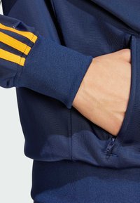 Marineblaue Sportjacke mit orangefarbenen drei Streifen an den Ärmeln, ausgestattet mit einer Reißverschlusstasche und gerippten Bündchen für einen figurbetonten Look.