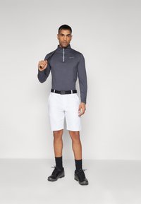 Szürke, hosszú ujjú póló fél cipzárral, fehér rövidnadrág, fekete öv és fekete sportcipő. A modell egy golfütőt tart. Egyszerű háttér.