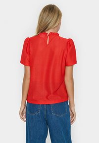 Blusa rossa a maniche corte con un motivo geometrico testurizzato, scollo arricciato e dettaglio a chiave sul retro, abbinata a jeans a gamba larga.