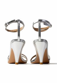 Paar silberne High-Heel-Sandalen mit Knöchelriemen, von hinten betrachtet, mit schlanken Absätzen und braunen Sohlen.