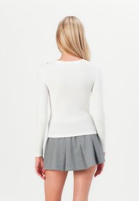 White long-sleeve top with a ribbed texture, featuring decorative hardware on the shoulder. Paired with a gray pleated skirt.