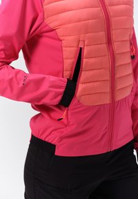 Lyserød sportjakke med quiltet koralfarvet frontpanel, lynlåslommer, elastiske manchetter og sorte detaljer ved taljen og lommer.