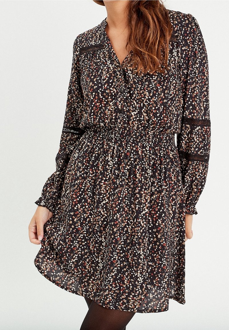 Femme portant une robe noire à manches longues avec des taches éparses marron, blanches et beiges, taille cintrée et détails en dentelle sur les manches.