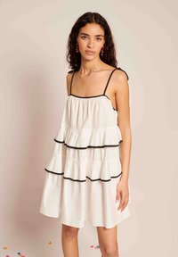 Vestido branco em camadas com acabamento preto, com alças finas nos ombros e camadas franzidas para dar textura. Modelo posa contra um fundo neutro.