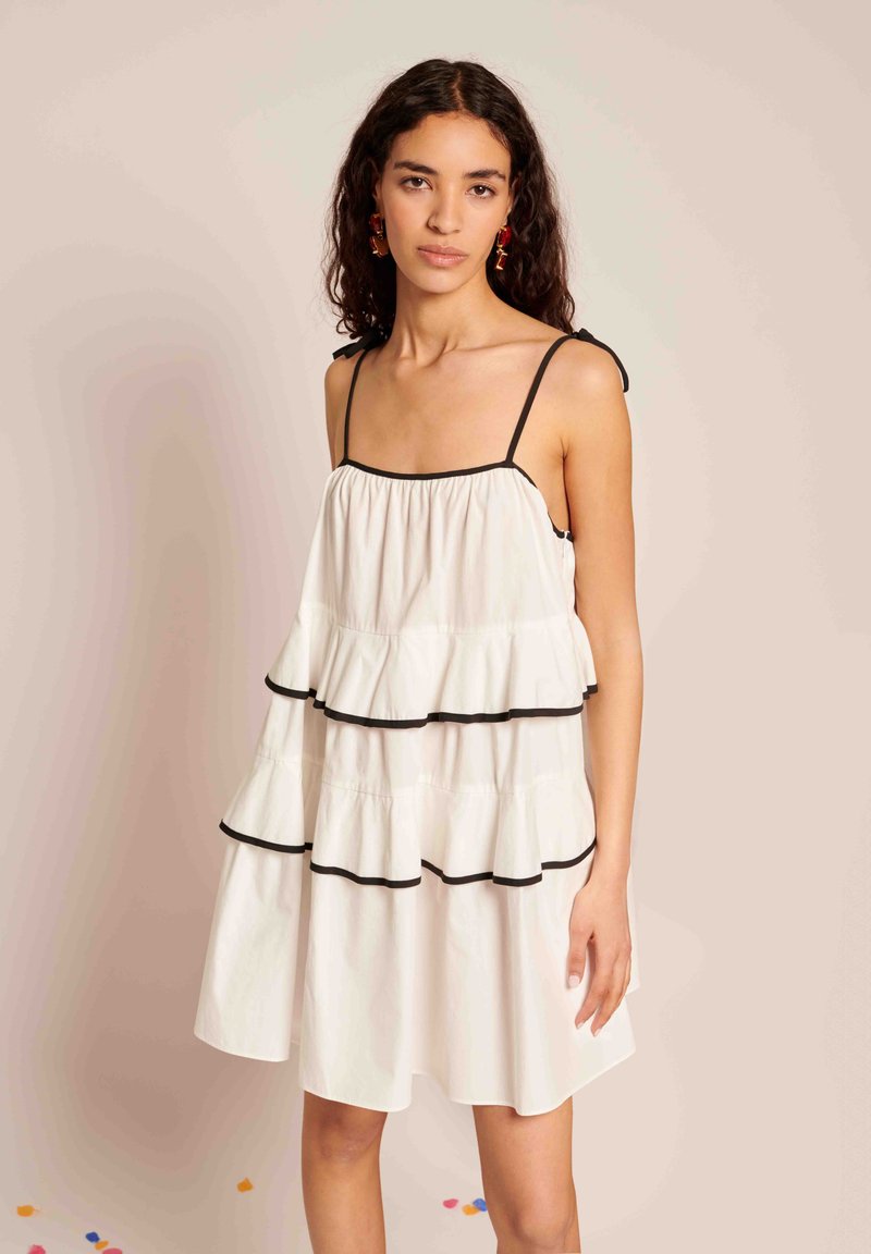 Vestido branco em camadas com acabamento preto, com alças finas nos ombros e camadas franzidas para dar textura. Modelo posa contra um fundo neutro.