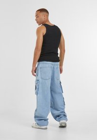Svart ermeløs tanktopp og lyseblå oversized cargo-bukser med sidelommer, tatt på sammen med hvite sneakers. Bakutsikt mot en lys bakgrunn.