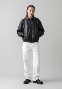 Ung person med vådt hår iført en sort læderjakke, hvide bukser og sorte sko, stående mod en ensfarvet lys grå baggrund.