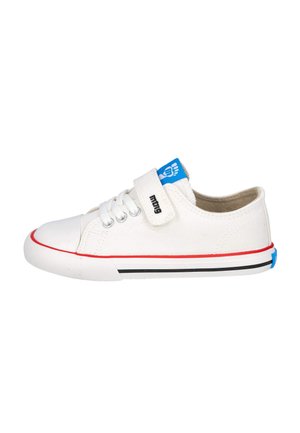 Zapatilla de lona blanca con una etiqueta azul, franja roja a lo largo de la suela, acentos en negro y blanco, y una correa de gancho y lazo junto a los cordones.