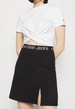 Woman wearing a white twisted crop top and a black skirt with a Calvin Klein Jeans waistband and front slit, standing against a plain background.