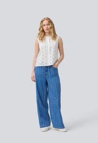 White sleeveless top with lace detailing, paired with loose blue striped denim trousers. Model stands with one hand in pocket.