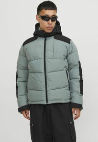 Padded jacket in muted green and black, featuring a zip closure, hood, and raised stitching. Textured fabric with a smooth finish.