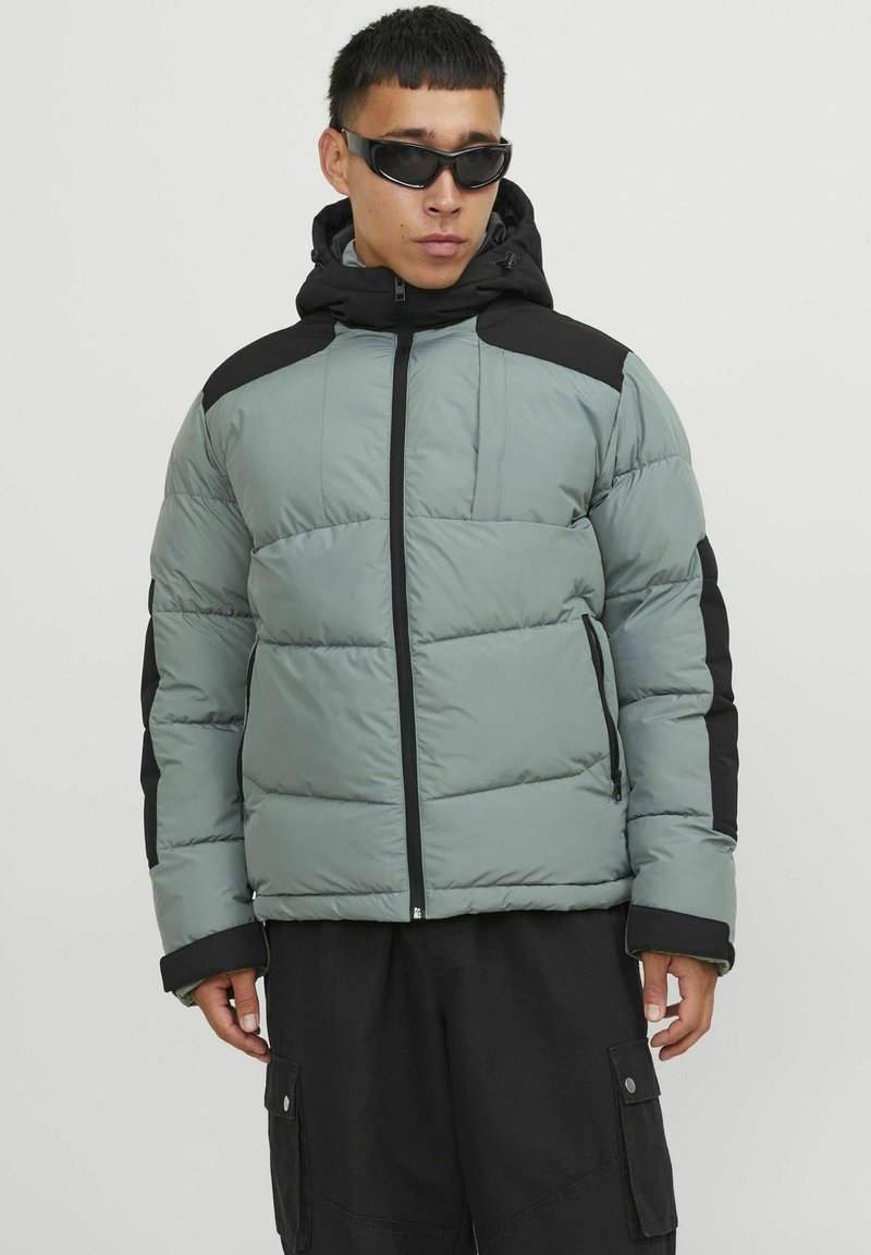 Padded jacket in muted green and black, featuring a zip closure, hood, and raised stitching. Textured fabric with a smooth finish.