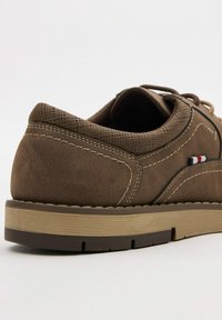 Zapato de cuero marrón con empeine texturizado, costuras en contraste y suela de goma color beige. Presenta un pequeño acento en rojo, blanco y azul.