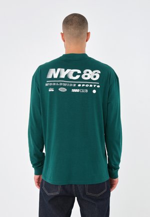 Camiseta de manga larga verde con "NYC 86" en blanco en la parte trasera, con logotipos y texto adicionales. Fabricada en tela suave, de corte relajado y diseño clásico.