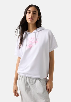 Vrouw met lang donker haar, gekleed in een witte korte mouw hoodie met roze afbeelding en lichtgrijze broek, staand tegen een effen achtergrond.