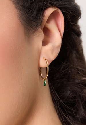 Złota kolczyka w kształcie obręczy z małym zielonym kamieniem jako zawieszką, gładka faktura i okrągły kształt, noszona na uchu modelki.