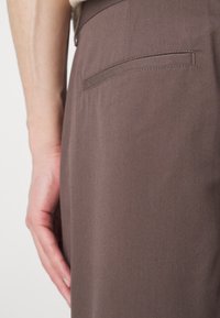 Bruine broek zijaanzicht, met een gladde textuur, riemlussen en een enkele achterzak met een gestikte flap.