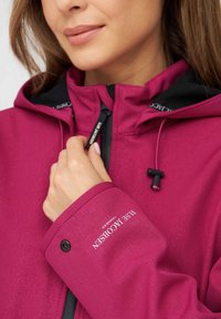 Chaqueta impermeable fucsia con acabado texturizado, capucha negra ajustable y tirador de cremallera grabado. Presenta un logo en el puño.