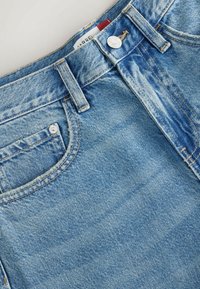 Gros plan sur un jean en denim bleu montrant la poche avant, la boucle de ceinture, le bouton métallique et une partie de la ceinture avec une petite étiquette en tissu.