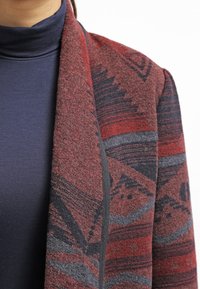 Personne portant un col roulé bleu marine sous une veste ou un châle à motifs rouges et gris, visible du cou jusqu'au milieu du torse.