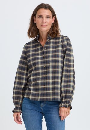 Kvinde med skulderlangt brunt hår iført blå, brun og gul ternet skjorte med knapper og blå jeans, stående mod en ensfarvet baggrund.