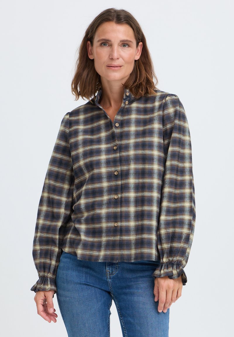 Femme aux cheveux bruns mi-longs portant une chemise à carreaux bleue, marron et jaune à boutons et un jean bleu, debout devant un fond uni.