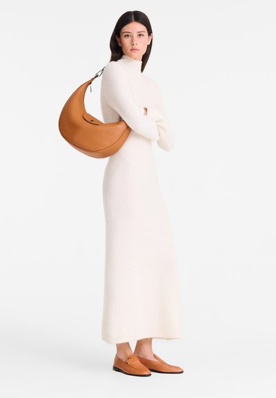 Woman wearing a long white knit dress, carrying a large orange leather shoulder bag, and matching orange loafers, standing against a white background.